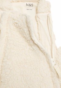 Giacca in pile color crema con una superficie strutturata e soffice. Presenta una zip frontale con tirante in tessuto e un'etichetta stampata all'interno.