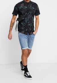 Maglietta nera a maniche corte con motivo di foglie verdi, pantaloni corti in denim blu chiaro, sneakers bianco e nere e una piccola borsa a tracolla nera.