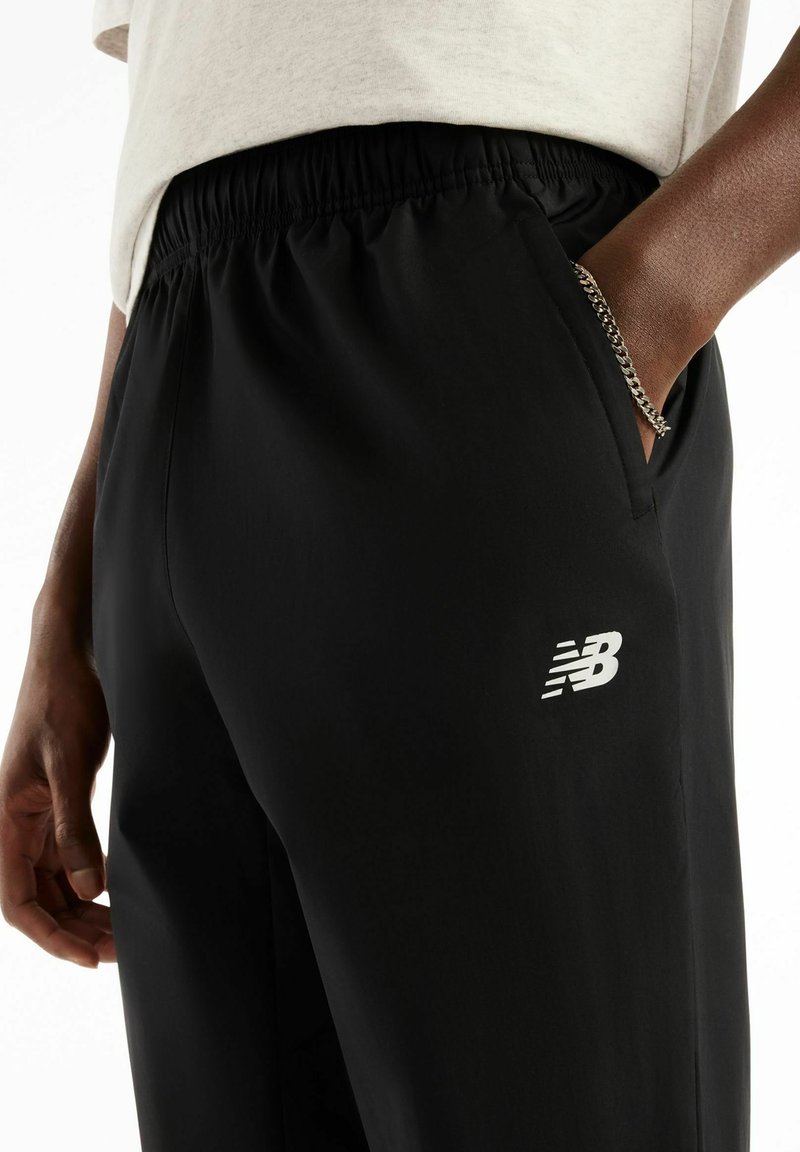 Person wearing black New Balance pants with elastic waist and hand in pocket, also wearing a silver chain bracelet.