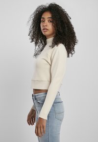 Beige kortärmad sweatshirt med långa ärmar och hög krage, tillsammans med ljusblå jeans, visad från sidan mot en neutral bakgrund.