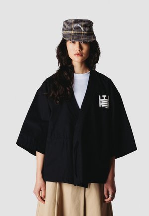 Vrouw die een zwart kimono-stijl jasje draagt over een wit overhemd, een beige rok en een geruite pet met lang donker golvend haar tegen een effen achtergrond.