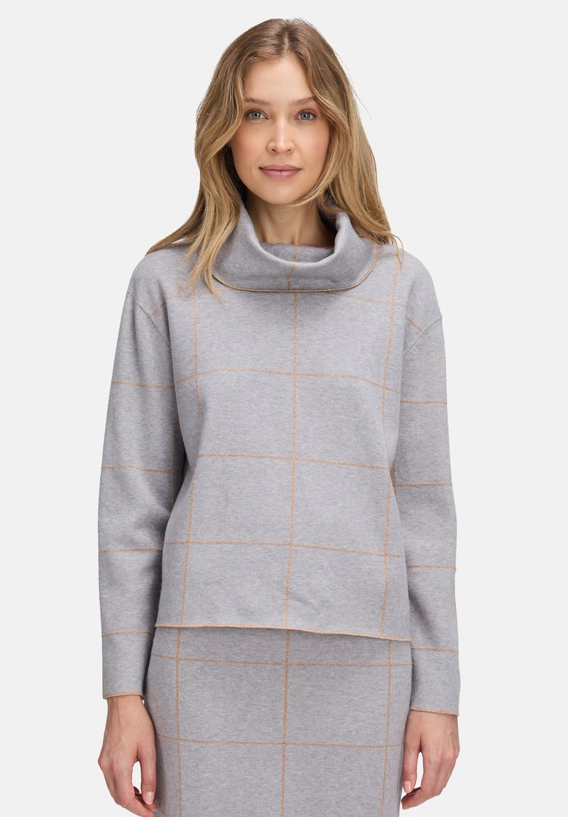 Kobieta wearing light gray knit top and skirt set with orange grid lines and a loose turtleneck collar, standing against a white background.