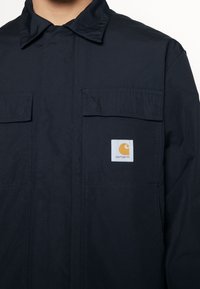 Navy blue work jacket made of durable fabric. Features a classic collar, two chest pockets, and a square logo patch on the left pocket.