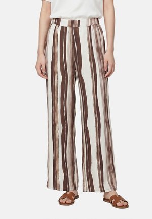 Personne portant un pantalon large à rayures verticales marron et beige, un haut blanc et des sandales marron à enfiler, debout devant un fond uni.