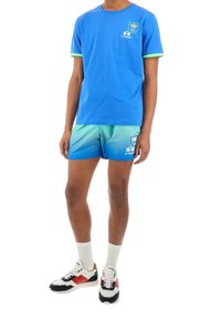 Camiseta de algodón azul con ribete verde, combinada con pantalones cortos degradados en verde y azul que tienen un logo. Calzado: zapatillas blancas con detalles en rojo y verde.