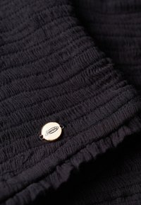 Sort tekstureret stof med vandret ribbing, der har et guld cirkulært logo-label med "Superdry" indgraveret. Blødt, fleksibelt materiale.