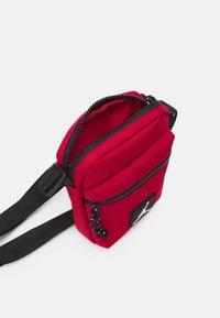 Borsa a tracolla rossa in tessuto con zip superiore, due scomparti, dettagli neri e una patch con logo. Superficie texturizzata con tracolla regolabile.
