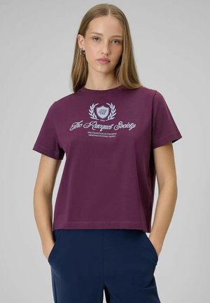 Młoda kobieta z prostymi włosami nosi fioletową koszulkę z napisem „The Racquet Society” oraz granatowe spodnie, ręce w kieszeniach, neutralne tło.