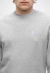 Mand iført en lysegrå crewneck-sweatshirt med et lille firkantet logo på venstre bryst mod en ensfarvet baggrund.