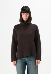 Femme avec des cheveux raides et foncés portant un pull à col roulé marron foncé et un jean bleu, se tenant devant un fond blanc uni.