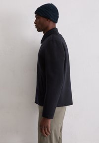 Granatowa marynarka z mieszanki wełny z wyraźnym kołnierzem, bocznymi kieszeniami i prostym dołem; zestawiona z jasnymi spodniami i dzianinową czapką.