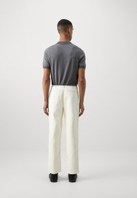 Tricou polo texturat gri cu mâneci scurte și guler, asociat cu pantaloni albi cu picioare drepte și pantofi negri. Fundal simplu neutru.