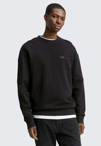 Sort sweatshirt med tekstureret overflade, der har en rund halsudskæring og et diskret logo-patch på brystet. Sammen med mørke jeans.