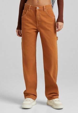 Femme portant un pantalon large marron taille haute avec poches avant et latérales, associé à des baskets blanches sur fond gris.