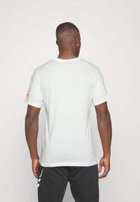 Biała bawełniana koszulka z krótkim rękawem, okrągłym dekoltem i małym czerwonym detalem na prawym rękawie, gładka tekstura, luźny krój.