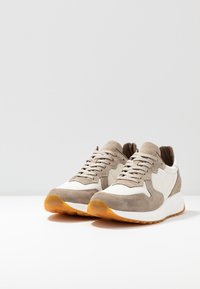 Beige ja valged tossud, millel on oranž kummist põhi, valgel pinnal heleda halli tausta ees.