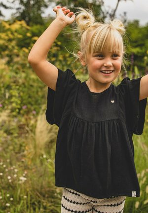 Zwarte top met korte mouwen en een geplooide voorkant met hartaccent, gecombineerd met gestreepte crèmekleurige en zwarte leggings in een natuurlijke omgeving.