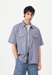 Camisa de cuadros de manga corta en azul, blanco y marrón, que cuenta con dos bolsillos frontales, cierre de botones y un corte relajado. Textura típica de tejido.