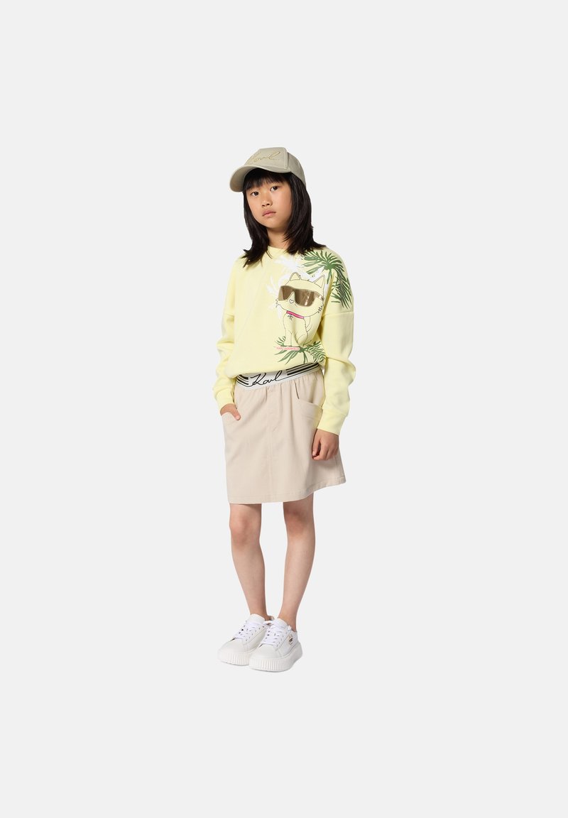 Sudadera amarilla con gráfico de gato y hojas de palma, combinada con una falda beige y zapatillas blancas. Gorra beige completa el conjunto.