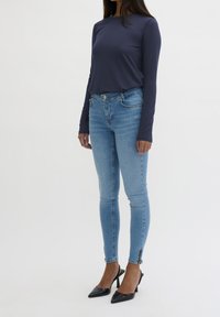Eine Frau trägt ein marineblaues Langarmoberteil, hellblaue enge Jeans mit Knöchelreißverschlüssen und schwarze spitze High Heels mit Slingback.