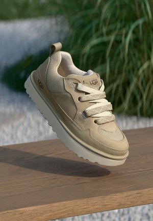 Beige sneakers van mesh en suede materiaal, met platte veters, een opvallend logo en een gestructureerde zool, tentoongesteld op een houten oppervlak.