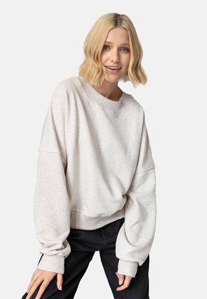 Blondynka o falowanych włosach nosi oversize'ową, jasnoszarą bluzę i czarne spodnie, lekko pochylona do przodu na jednolitym tle.