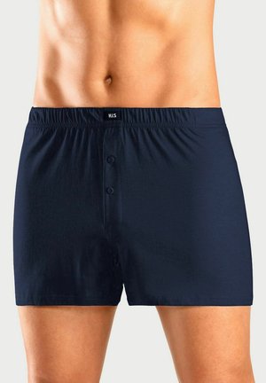 Male torso wearing navy blue boxer shorts with front buttons and elastic waistband, standing against a plain light background.