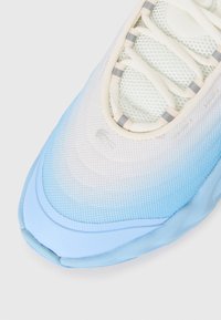 Gros plan sur une sneaker bleue et blanche avec tissu en maille et boucles de lacet réfléchissantes sur fond blanc.