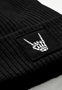 Black knit beanie featuring a woven black patch with a white skeleton hand gesture. The fabric has a textured, ribbed appearance.