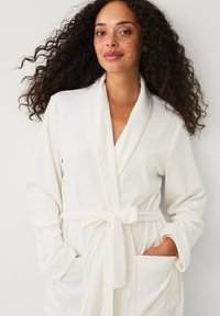 Vrouw met krullend haar en sproetjes, gekleed in een witte badjas die in de taille is gestrikt, met handen in de voorzakken, staand tegen een effen achtergrond.