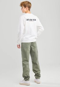 Camisa blanca de manga larga con estampado "Into the Wild" en la parte posterior; combinada con pantalones cargo verde claro y zapatillas blancas. Un atuendo sencillo y casual.