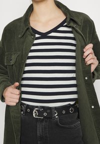 Khaki korduroy košile přes černobíle pruhovanou V-krkovou košili, s černým páskem se stříbrnými kroužkovými okují, spárované s tmavými džíny.