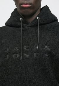 Κοντινό πλάνο ενός ατόμου που φορά μαύρη ανάγλυφη φούτερ με κουκούλα, με το "JACK & JONES" κεντημένο στο στήθος και τα κορδόνια να κρέμονται από την κουκούλα.