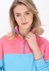 Sudadera de media cremallera en rosa y azul, con una textura suave y lisa. Presenta mangas en contraste y una cremallera con un tirador circular.