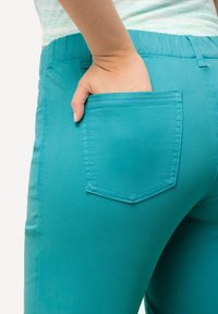 Pantaloni turchesi con una texture liscia, dotati di una tasca posteriore di forma rettangolare. Vita elastica visibile nella parte superiore.