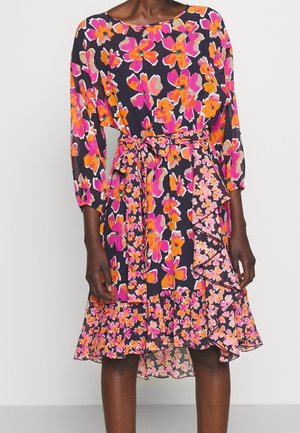 Kvinde iført en knælængde sort kjole med lyserød, orange og beige blomsterprint og et matchende stofbælte bundet om taljen.