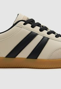 Zapatilla beige con rayas negras, suela de goma y acentos texturizados. Presenta cordones negros y detalles de costura en un material de cuero suave.