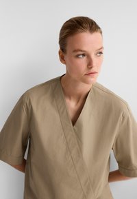 Beige kurzärmlige Wickelbluse aus leichtem Stoff, mit V-Ausschnitt und lockerer Passform. Minimalistisches Design ohne sichtbare Muster.