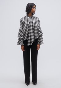 Μπλούζα με ασπρόμαυρο καρό σχέδιο, με βολάν στα μανίκια, ψηλό λαιμό και ασύμμετρο τελείωμα. Συνδυασμένη με μαύρο παντελόνι ίσιας γραμμής και ψηλοτάκουνα.