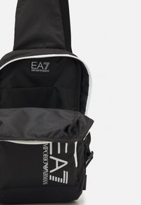 Borsa a tracolla nera in nylon, con una zip bianca, logo sul davanti e un ampio scomparto principale con foderatura morbida.