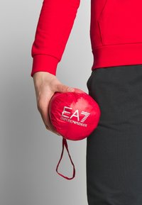 Czerwona kompaktowa saszetka z logo "EA7 EMPORIO ARMANI". Gładka tekstura, okrągły kształt i pasek do noszenia. Trzymana w dłoni, z widoczną czarną odzieżą.