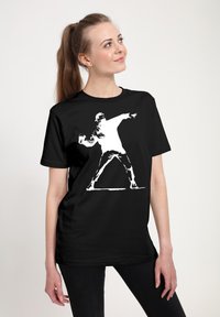Mladá žena s copem, oblečená v černém tričku s bílou postavou ve stylu šablony házející předmět, stojící na jednobarevném pozadí.
