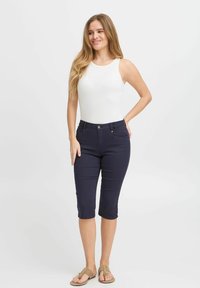 Pantaloni capri blu navy con un design aderente, tasche frontali e dettagli di cucitura sottili, abbinati a un top senza maniche bianco. I sandali sono beige.
