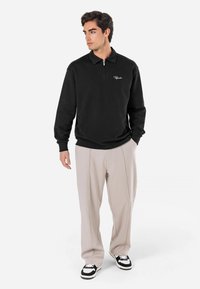 Sort zip-up sweatshirt med krave, lange ærmer og et hvidt broderet logo på brystet, parret med beige bukser og sorte sneakers.