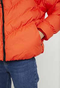 Bright orange puffer jacket with diagonal stitching, zippered front, and elastic cuffs, worn over blue jeans. Textured finish visible.