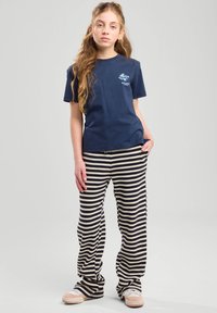 T-shirt di cotone navy con stampa azzurra chiara, abbinato a pantaloni larghi a righe nere e crema. Il modello indossa delle scarpe beige slip-on.