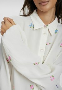 Blusa bianca con ricami floreali in rosa e blu, dotata di colletto, chiusura con bottoni e maniche lunghe e vaporose. Tessuto morbido e strutturato.