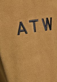 Brauner Fleece-Stoff mit weicher Textur, der die gestickten Initialen "ATW" in Schwarz zeigt.