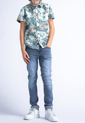 Camicia floreale a maniche corte in teal e bianco, jeans blu e scarpe da ginnastica bianche. La camicia presenta grandi motivi a foglia e una chiusura con bottoni.