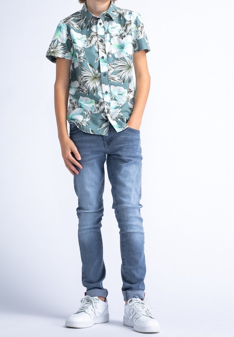 Camicia floreale a maniche corte in teal e bianco, jeans blu e scarpe da ginnastica bianche. La camicia presenta grandi motivi a foglia e una chiusura con bottoni.